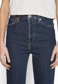 Jeans in denim blu scuro con vita alta, dotati di rivetti in rame, chiusura con bottone frontale e senza motivi o segni di usura visibili.