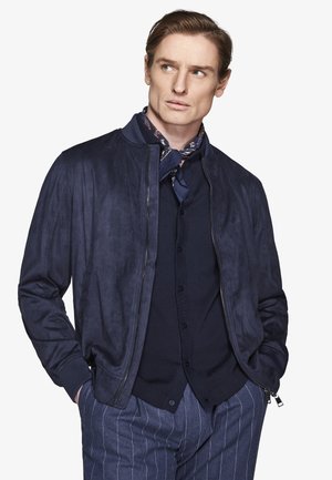 Mężczyzna w granatowej bomberce zamszowej, ciemnej koszuli zapiętej na guziki, wzorzystym szaliku na szyi i niebieskich spodniach w prążki, z rękami w kieszeniach, patrzący na bok.