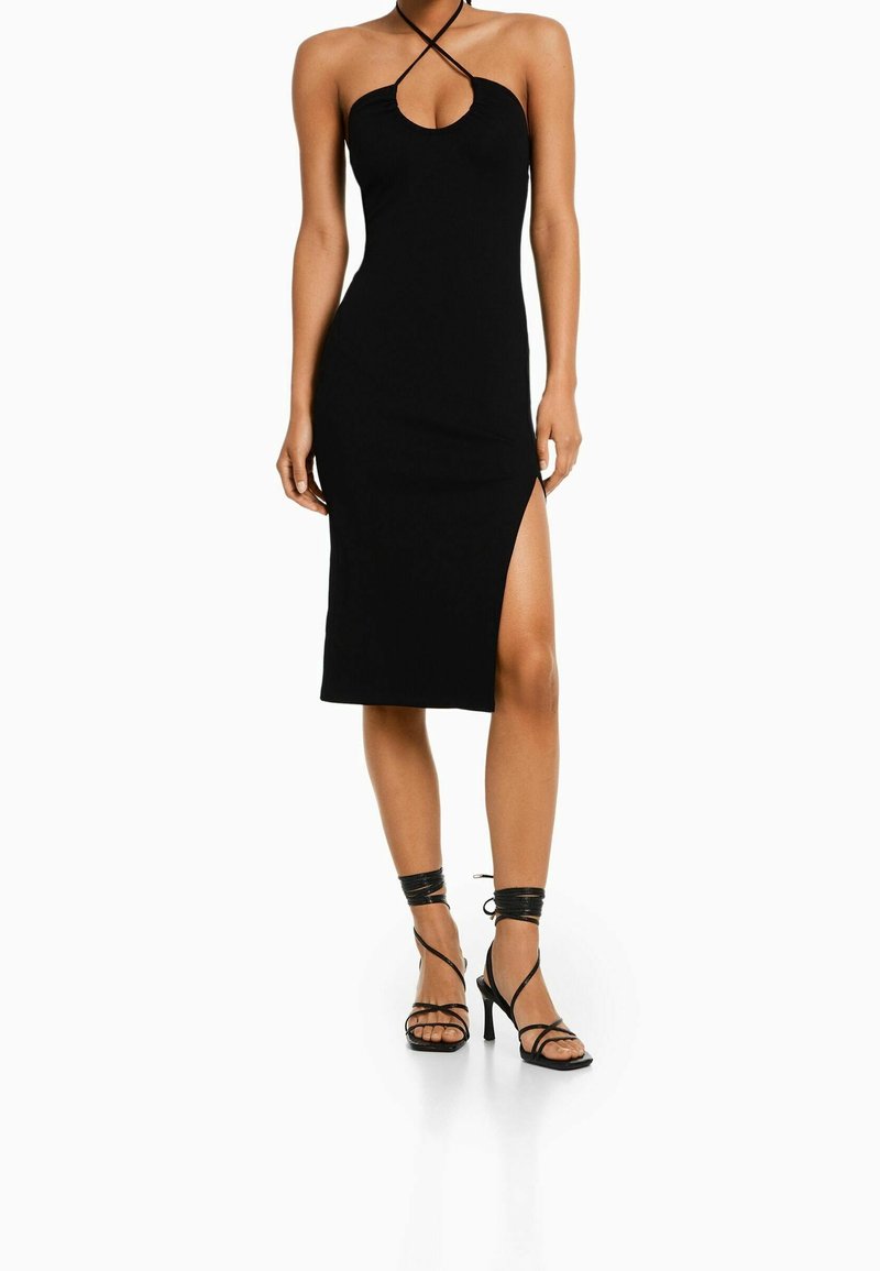 Femme portant une robe midi noire ajustée avec des bretelles croisées au cou, une découpe à l'avant, une fente sur la cuisse, et des sandales à talons hauts noires à lacets.
