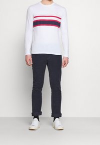 Weißes Langarmhemd mit roten und marineblauen horizontalen Streifen, kombiniert mit marineblauen Hosen und weißen Turnschuhen. Einfaches Design und figurbetonte Passform.