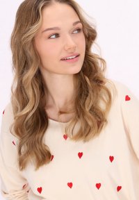 Maglione chiaro con motivi a cuori rossi sparsi, caratterizzato da un collo ampio e capelli ondulati parzialmente visibili su uno sfondo neutro.
