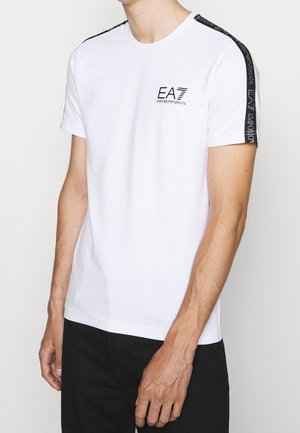 Mężczyzna w białej koszulce z krótkim rękawem z czarnymi, oznaczonymi paskami na ramionach i logo "EA7 Emporio Armani" na piersi.