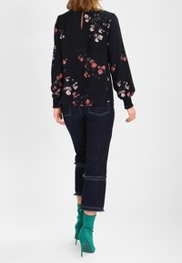 Blusa nera a maniche lunghe con stampa floreale, schiena a chiave, e polsini elastici, abbinata a pantaloni cropped in denim scuro con orli sfrangiati e tacchi verdi.