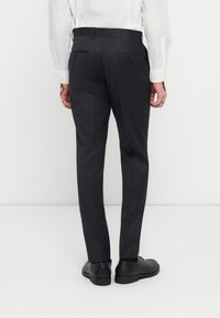 Dunkelgraue, maßgeschneiderte Hose mit schlanker Passform, ausgestattet mit zwei Gesäßtaschen mit Knopfverschlüssen, kombiniert mit schwarzen Schuhen.