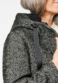 Gestructureerde zwart-wit herringbone-patroonjas met capuchon, met een accent van zwarte lint, te zien nabij de schouder van een persoon.