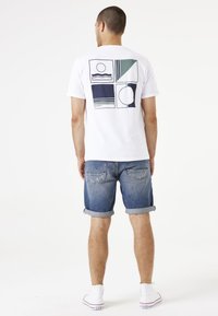 Camiseta blanca con un diseño gráfico geométrico en verde y azul en la parte posterior, combinada con pantalones cortos de mezclilla enrollados y zapatillas blancas.