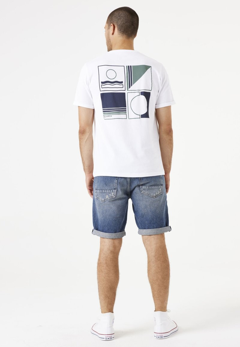 Camiseta blanca con un diseño gráfico geométrico en verde y azul en la parte posterior, combinada con pantalones cortos de mezclilla enrollados y zapatillas blancas.