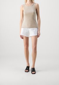 Kvinna iklädd en beige ärmlös stickad topp, vita fransiga denimshorts och svarta öppna klackskor, stående mot en enkel ljus bakgrund.