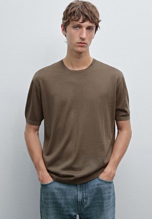 Brown knitted T-shirt with a round neckline and short sleeves, paired with light blue denim jeans, on a model against a plain backdrop.