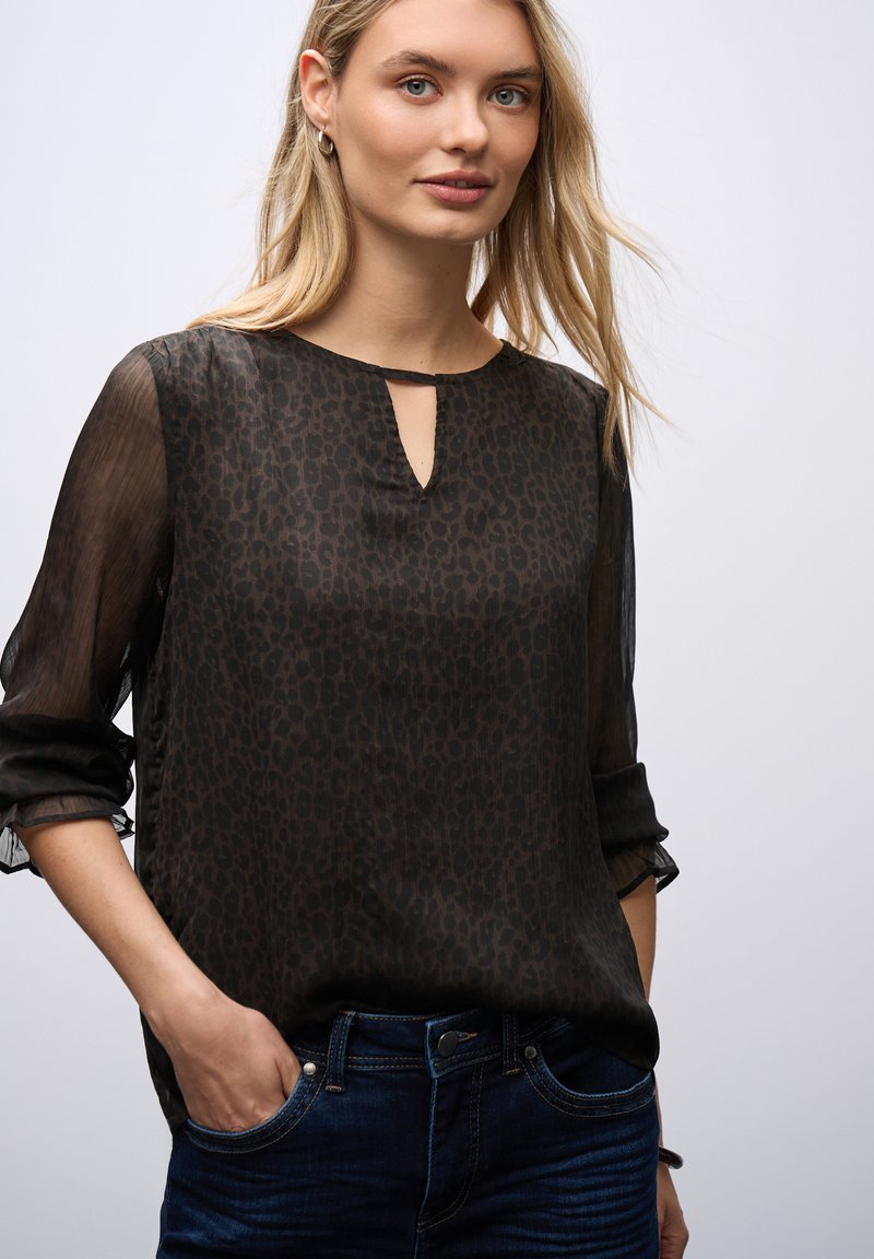Blusa nera trasparente con motivo a stampa leopardo, scollatura a chiave e maniche a tre quarti. Abbinata a jeans blu scuro.
