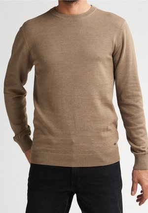 Man wearing a light brown knit sweater with long sleeves and ribbed cuffs, paired with black pants, standing against a plain background.