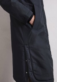 Schwarze gepolsterte Jacke mit einer Seitentasche, Druckknöpfen am Saum und einer glatten Textur. Sie hat eine elegante, taillierte Silhouette.