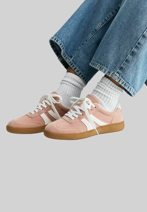 Person trägt hellrosa Wildledersneaker mit weißen Streifen und Schnürsenkeln, weiße gerippte Socken und blaue Jeans vor einfarbigem Hintergrund.