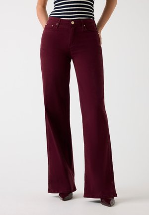 Donna con pantaloni a vita alta bordeaux scuro a gamba larga abbinati a un top senza maniche a righe bianche e nere, in piedi con le mani in tasca.
