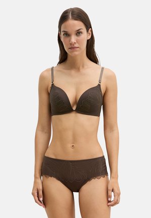 Frau mit langen braunen Haaren, die ein passendes braunes BH- und Slip-Set mit floraler Stickerei trägt, steht vor einem einfarbigen hellen Hintergrund.