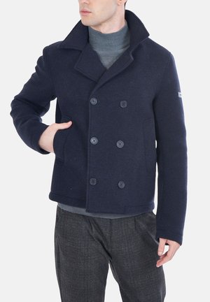 Uomo che indossa un cappotto doppio petto blu navy con collo rialzato sopra un dolcevita grigio e pantaloni scuri a quadri.