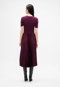 Dlhé plisované šaty v tmavej burgundskej farbe, s priliehavým rebrovaným korzetom a krátkymi rukávmi, skombinované s čiernymi čižmami po kolená.