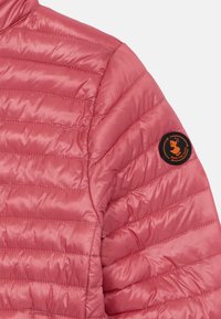 Light pink puffer jacket with quilted design, featuring a black patch on the shoulder with an orange logo. Smooth, shiny texture.