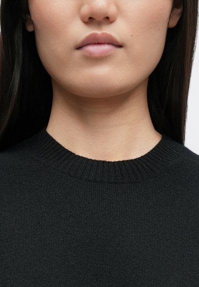 Μαύρο πουλόβερ πλεκτό με ριχτό στρογγυλό λαιμό, μαλακή υφή και εφαρμοστή σχεδίαση, σε ουδέτερο φόντο.