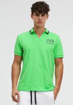 Junger Mann mit lockigem Haar, der ein leuchtend grünes Polohemd mit schwarzem Text und weiße Shorts vor einem einfarbigen Hintergrund trägt.