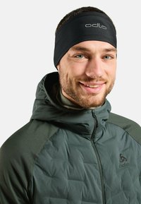 Bandeau de performance noir en tissu extensible, avec un logo. Porté avec une veste matelassée verte foncée à col.