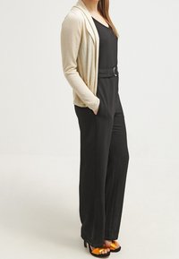 Beige kardi med avslappnad passform, parat med en svart jumpsuit som har ett bälte. Orange sandaletter med remmar fulländar outfiten. Släta texturer.