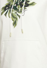 Felpa bianca con design di foglia di palma verde e testo nero "The BOSS", dotata di tasca frontale. I lacci hanno punte in metallo dorato.