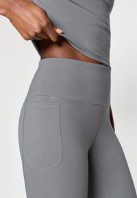Sive tajice s visokim strukom napravljene od rastezljivog materijala, s bočnim džepom i suptilnim detaljem šivanja. Glatka tekstura s pripijenim krojem.