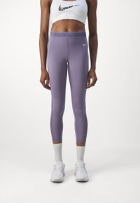 Fioletowe legginsy z elastycznej tkaniny z wysokim stanem, z napisem "NIKE PRO". Nosi je dziewczyna w białym, krótkim topie sportowym i białych butach.