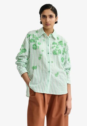 Donna con capelli corti che indossa una camicia a righe verde chiaro e bianca con ricamo floreale verde e pantaloni marroni a gamba larga, in piedi con una mano in tasca.