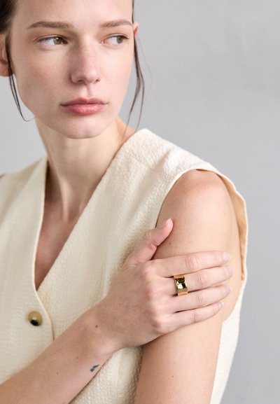 Bague en or avec une finition polie portée à la main, associée à un haut sans manches crème texturé avec un col en V et des boutons.