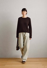 Pull en tricot noir, pantalon large vert clair, chaussures marron, tenant un sac noir, se tenant sur un sol en bois contre un mur gris.
