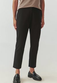 Čierne na mieru šité nohavice so slim fit strihom, po členky, s bočnými vreckami a hladkou textúrou. Nosia sa s tmavými, robustnými mokasínami.