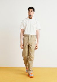 White short-sleeve t-shirt, beige straight-leg pants, and gray-orange sneakers. Model stands on a yellow floor with a plain background.