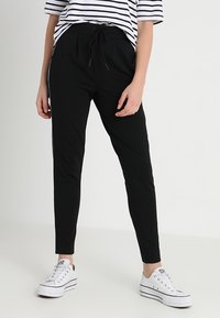 Calças de jogging pretas com cordão na cintura e riscas brancas laterais, feitas de um material liso e elástico, combinadas com ténis brancos.