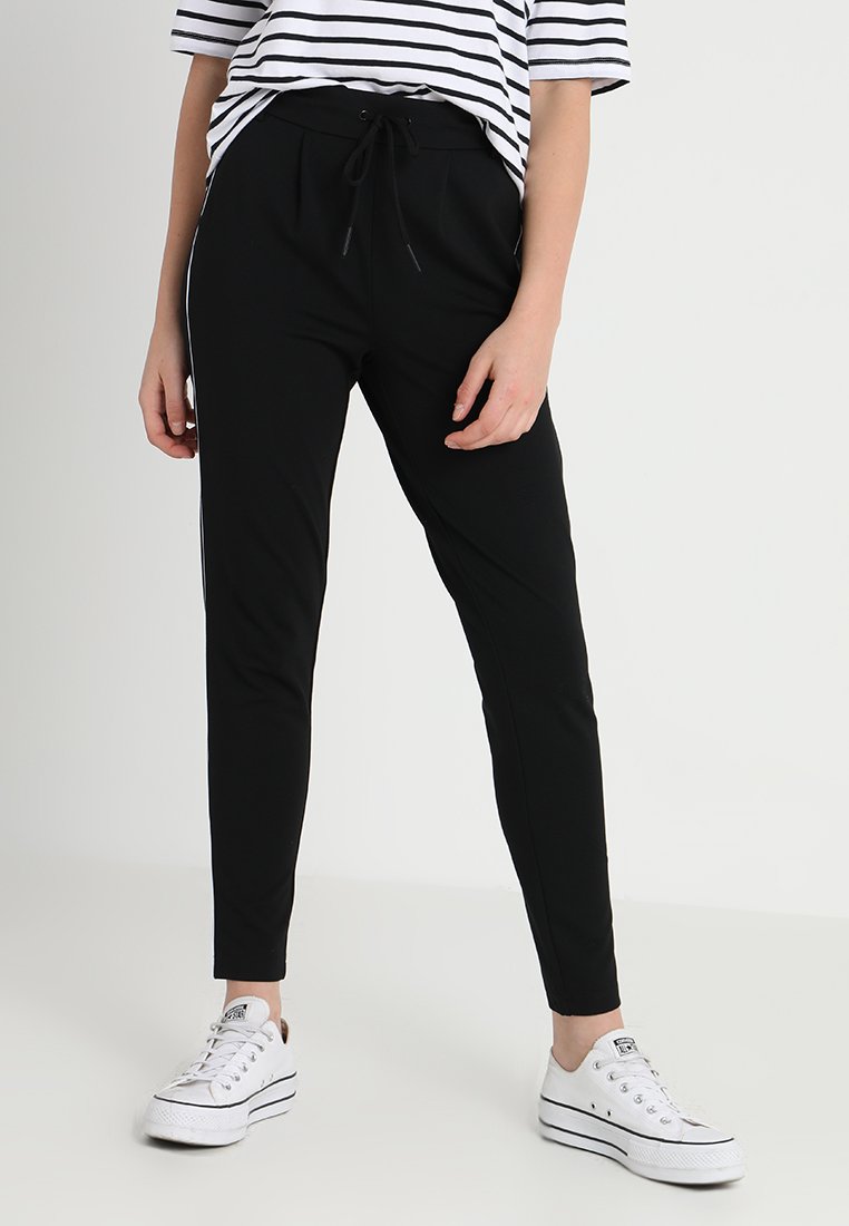 Calças de jogging pretas com cordão na cintura e riscas brancas laterais, feitas de um material liso e elástico, combinadas com ténis brancos.