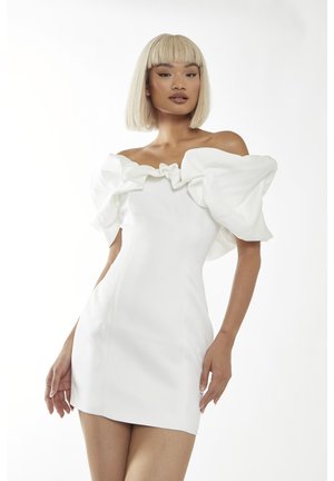 Frau mit kurzen blonden Haaren, die ein weißes Minikleid mit Carmenausschnitt und Puffärmeln vor einem schlichten weißen Hintergrund trägt.