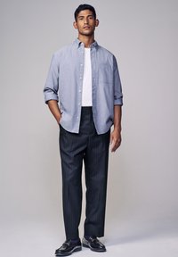 Camicia azzurra a maniche lunghe con colletto botton-down sopra una t-shirt bianca, abbinata a pantaloni scuri a righe e mocassini neri. Design semplice, vestibilità su misura.