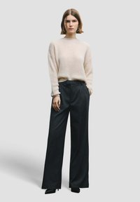 Pull beige en maille avec manches larges associé à un pantalon taille haute à fines rayures foncées. Le mannequin porte des escarpins noirs ajustés.