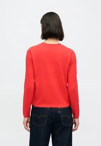 Suéter rojo de manga larga, con cuello redondo y textura suave. Combinado con jeans de mezclilla oscuros que presentan un diseño de bolsillo en contraste.