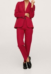 Completo rosso su misura con blazer aderente e pantaloni affusolati, abbinato a mocassini neri. Texture liscia e dettagli di design eleganti.