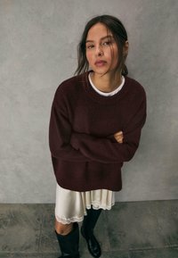 Bordeaux oversized sweater med ribbet halsudskæring, lagdelt over en cremet satin kjole med blondekant, parret med sorte knæhøje støvler.
