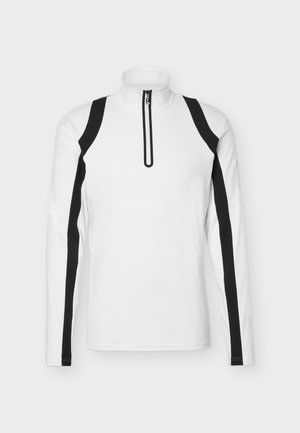 Joven de pie vistiendo una camiseta blanca de manga larga con cremallera y rayas negras, pantalones cortos negros, calcetines gris claro y zapatillas blancas sobre un fondo liso.