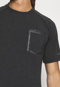 Mörkgrå t-shirt med kort ärm, tillverkad av ett texturerat material. Den har en kontrasterande, matt svart rektangulär ficka på vänster sida.