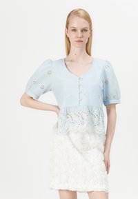 Blusa azzurra chiara con maniche a sbuffo corte, motivo ad occhiello e chiusura a bottoni; abbinata a una gonna bianca testurizzata con applicazioni floreali.