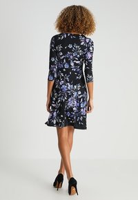Robe noire à fleurs avec des manches trois-quarts, ornée de fleurs violettes et blanches, jupe évasée et tissu léger. Escarpins noirs.
