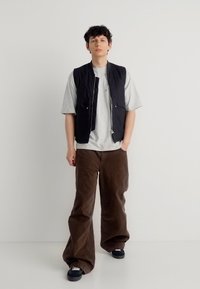 Νεαρός άνδρας που στέκεται φορώντας ένα μαύρο γιλέκο πάνω από γκρι μπλουζάκι, φαρδιά καφέ παντελόνια και μαύρα αθλητικά παπούτσια, σε απλό λευκό φόντο.