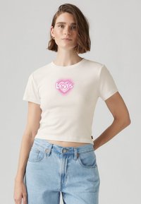 Krémové zkrácené tričko s krátkým rukávem, v centru se nachází růžové srdíčkové logo "Levi's". Měkká bavlněná tkanina, volný střih.