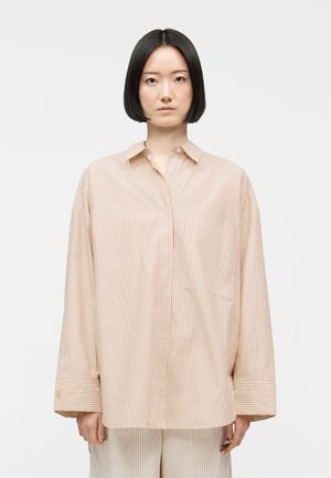 Žena s rovnými čiernymi vlasmi strihanými na bob nosí veľkú béžovo-bielu košeľu s vertikálnymi pruhmi a zodpovedajúce nohavice na bielom pozadí.