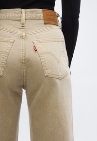 Μπεζ τζιν Levi's με δερμάτινο λογότυπο και κόκκινη ετικέτα, φορεμένα με μαύρο μακρυμάνικο τοπ, εμφανιζόμενα από πίσω στο ύψος της μέσης.